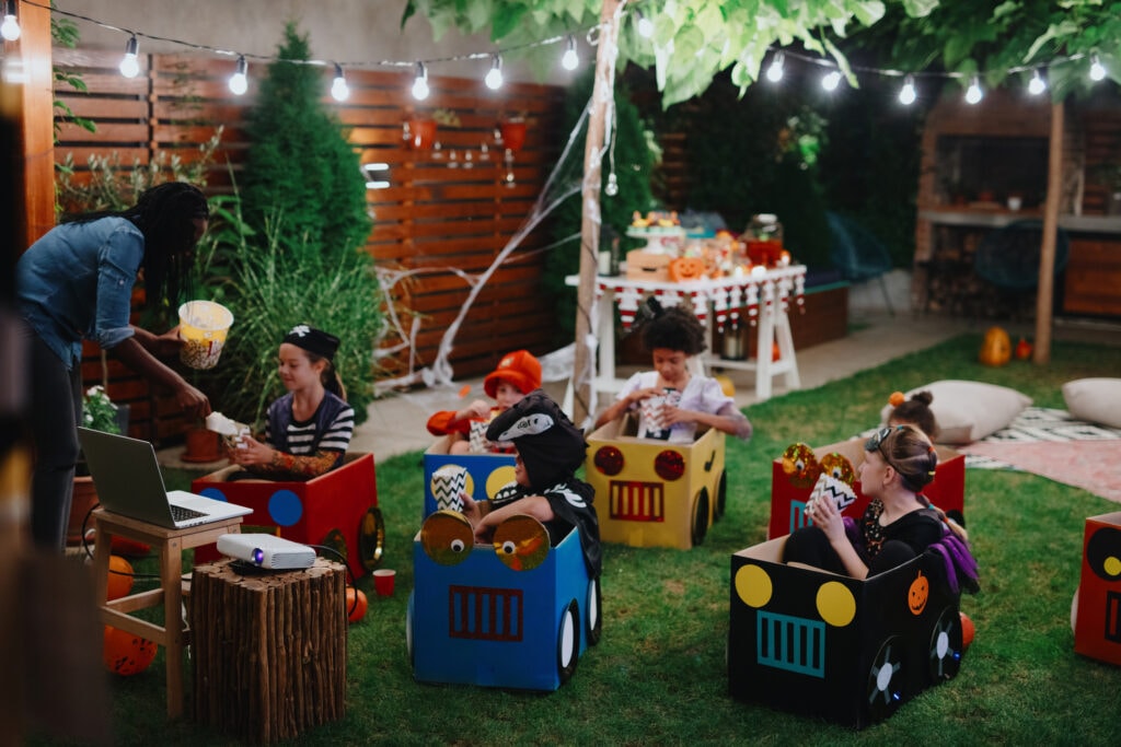 A mother serves popcorn to a group of kids in costumes, sitting in seats decorated like cars and trucks