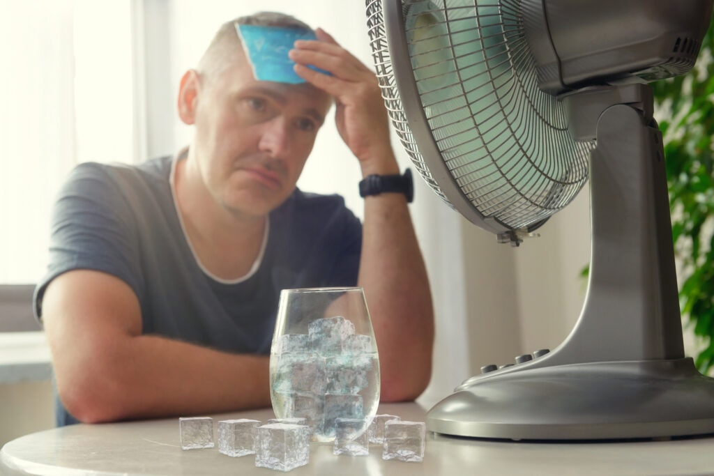 A man sitting in front of a table fan with cup of ice water