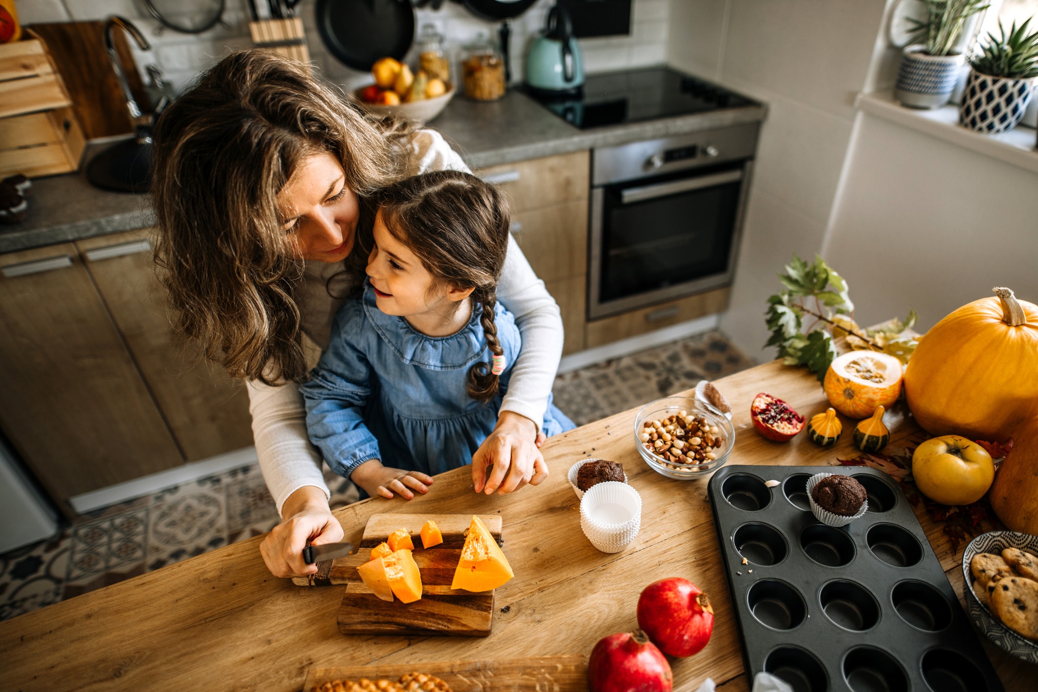 Person and child preparing food together in a kitchen, surrounded by pumpkins, fruit, and baking ingredients on a wooden countertop.