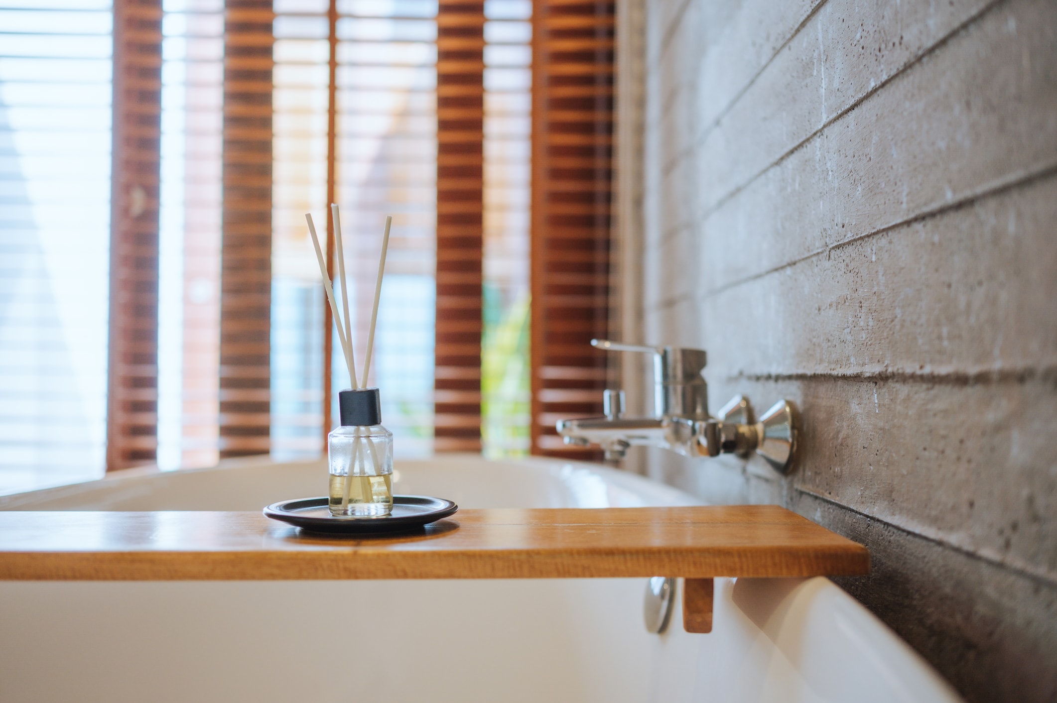 Modern bathroom with a freestanding bathtub, wooden bath tray, and reed diffuser beside a textured stone wall.