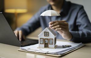 Small model house on documents with a hand holding an umbrella above it beside a laptop.