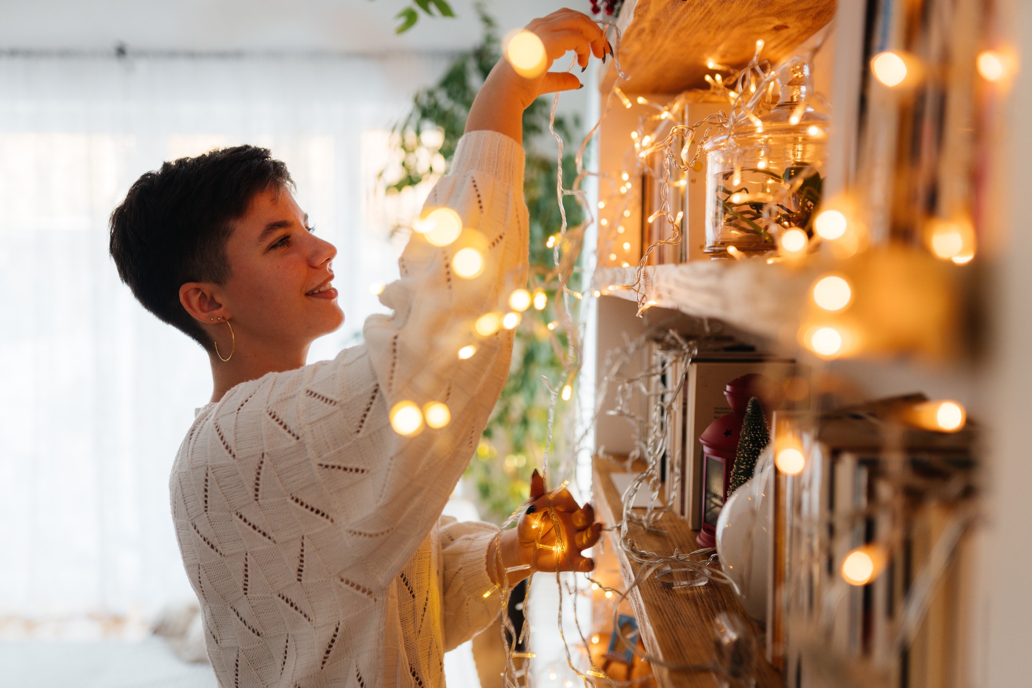 Person decorating a shelf with string lights in a cozy home setting, surrounded by warm ambient lighting and seasonal décor.