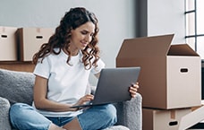 Person sitting on a couch using a laptop and tablet, with moving boxes in the background.