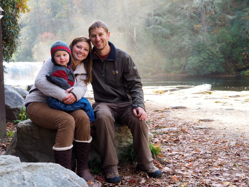 Family sitting on rocks beside a river in a wooded outdoor setting.