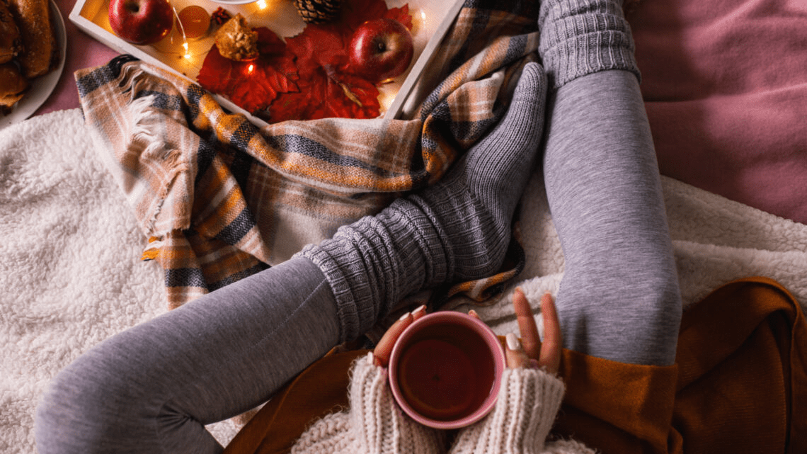 Person relaxing under a blanket with a warm drink, surrounded by cozy autumn décor and soft lighting.