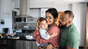 Personne tenant un jeune enfant dans ses bras pendant qu’une autre personne les enlace par derrière dans une cuisine lumineuse et accueillante.