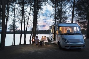 A group dining outdoors beside a camper van by a lakeshore at dusk.