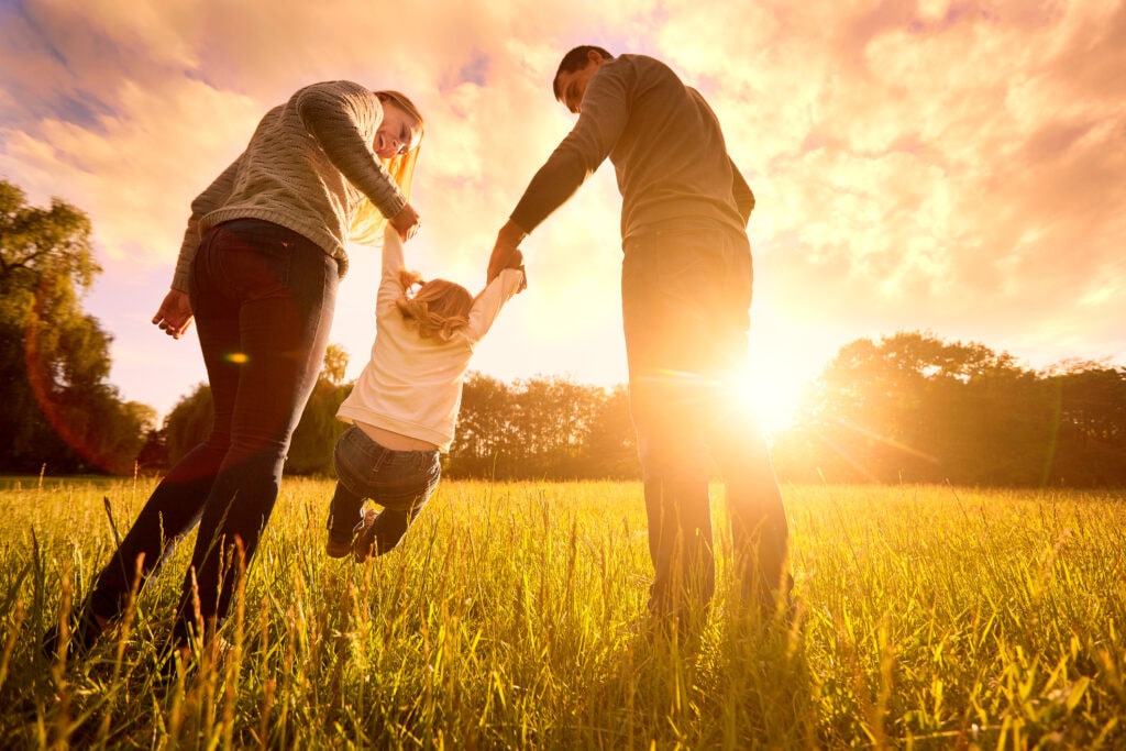 A young girl holds hands with her parents and swings in the air at sunset