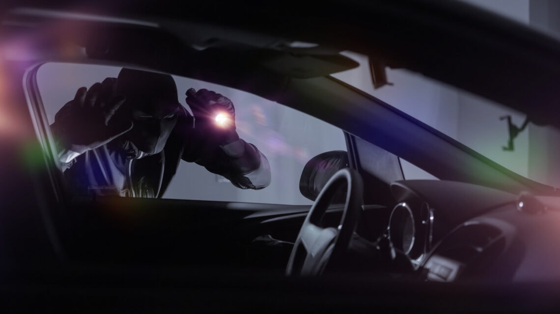 A car thief holds a flashlight against the window of a parked vehicle