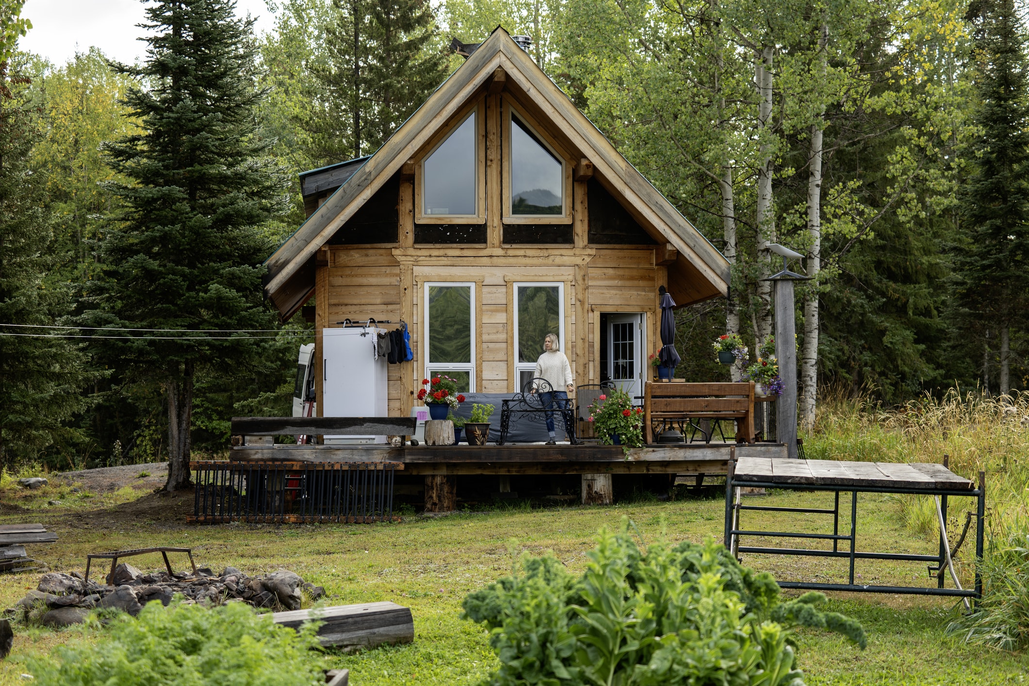 Wooden cabin with a deck surrounded by trees and a garden.