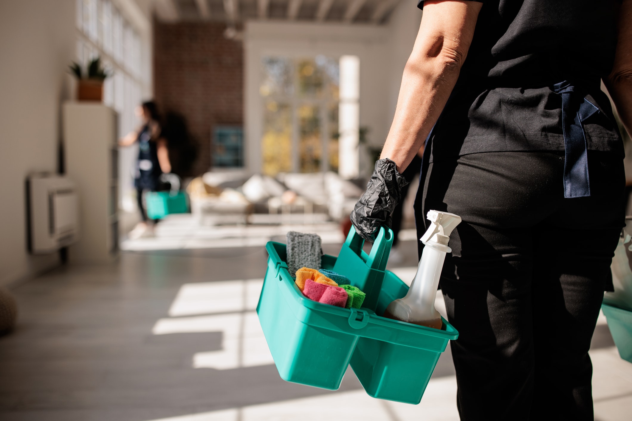 Person holding a cleaning caddy with spray bottle and cloths in a bright living space.