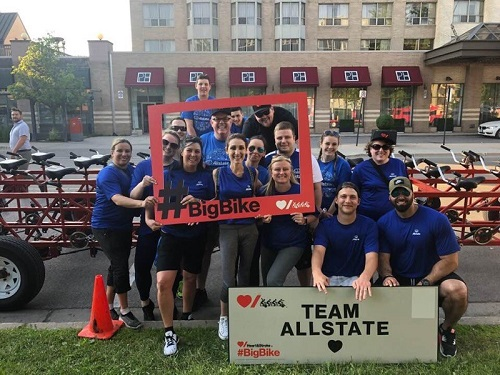 a group of Allstate employees in blue shirts holding a big bike and Team Allstate signs